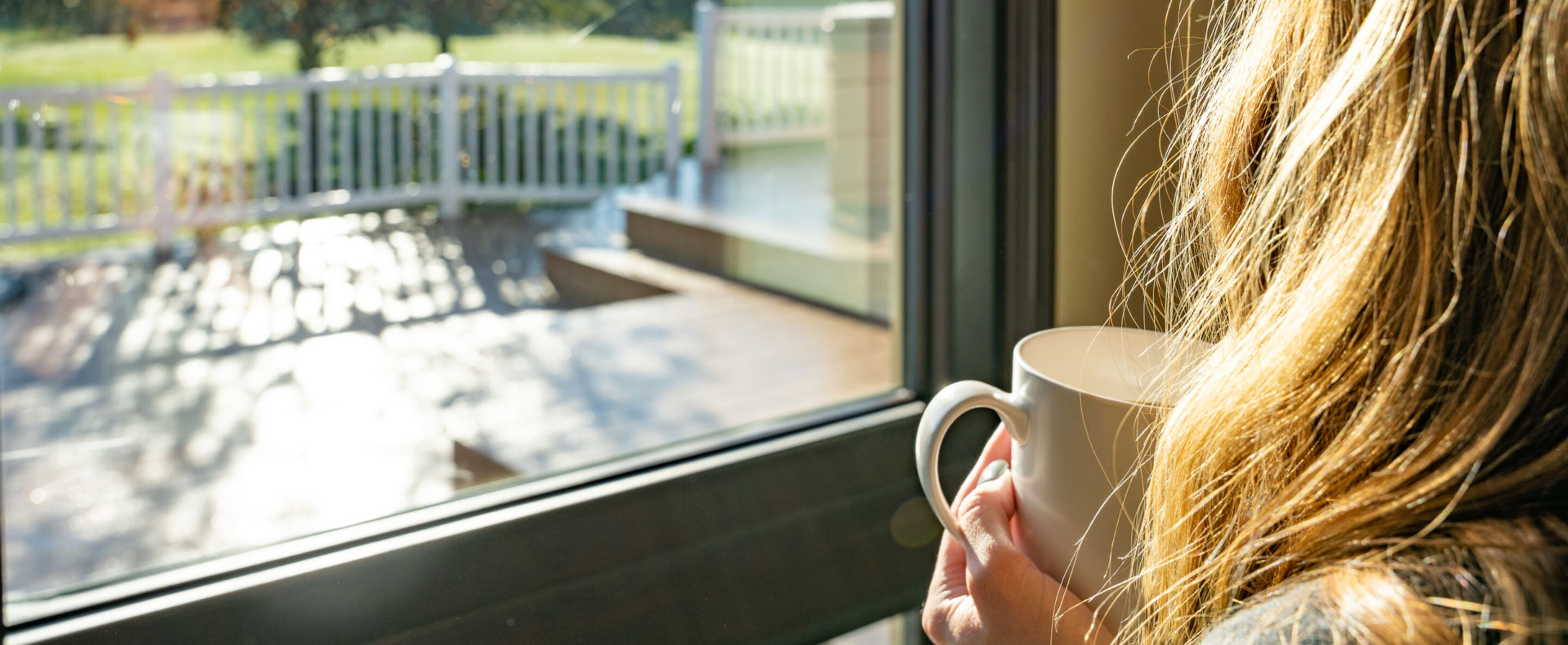 Woman holding a cup of coffee gazing out ProVia home replacement windows into her backyard, one of many vinyl replacement window types offered by ProVia