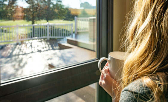 Woman holding a cup of coffee gazing out ProVia home replacement windows into her backyard, one of many vinyl replacement window types offered by ProVia