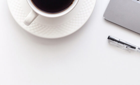 A cup of coffee on a white saucer next to a laptop, pen, notebook, and glasses with a hint of greenery of the far left corner