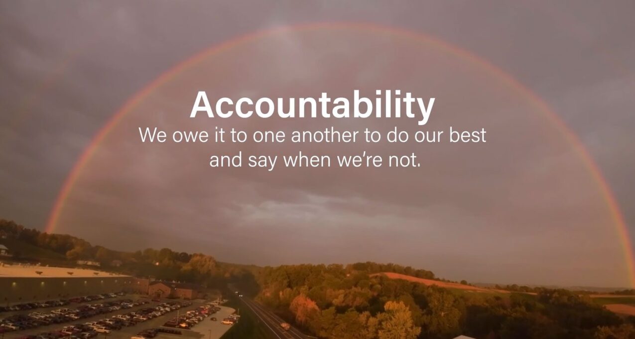 Image of a rainbow over some trees, a highway, and a parking lot with the words: 